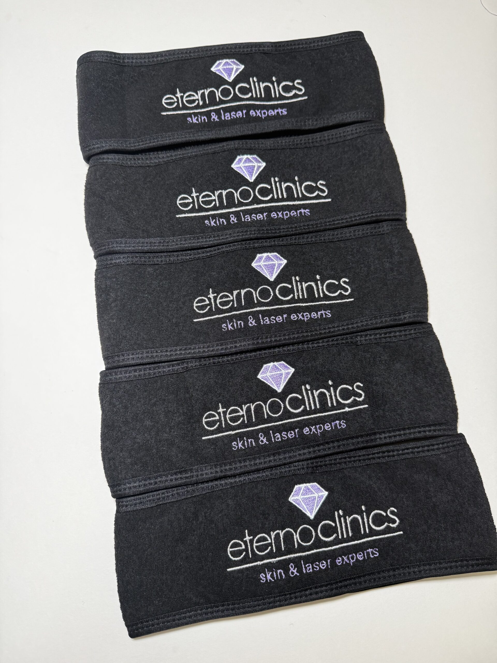 Black spa headbands embroidered with clinic logo in white thread, stacked neatly to show professional beauty salon branding for skincare and laser treatments.