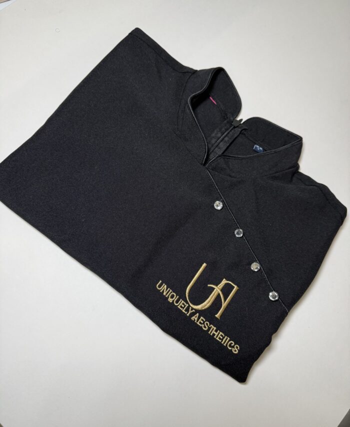 Black beauty therapist tunic with gold embroidered logo reading “Uniquely Aesthetics”, professional spa and aesthetic clinic uniform with custom logo embroidery, personalised workwear for salons, beauty therapists and medical aesthetic professionals.