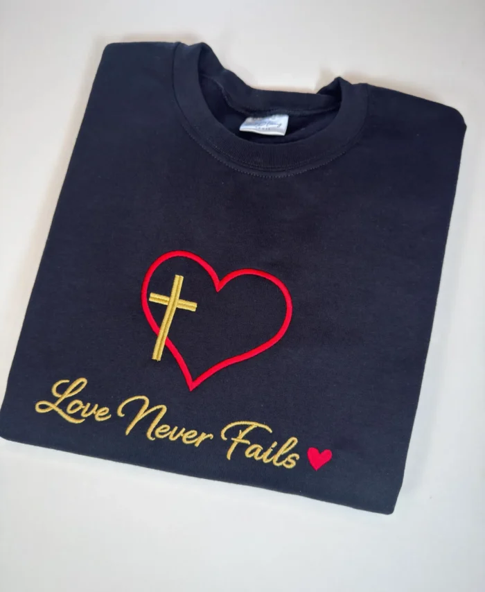 Black embroidered Christian T-shirt featuring red heart outline with gold cross and “Love Never Fails” Bible verse design