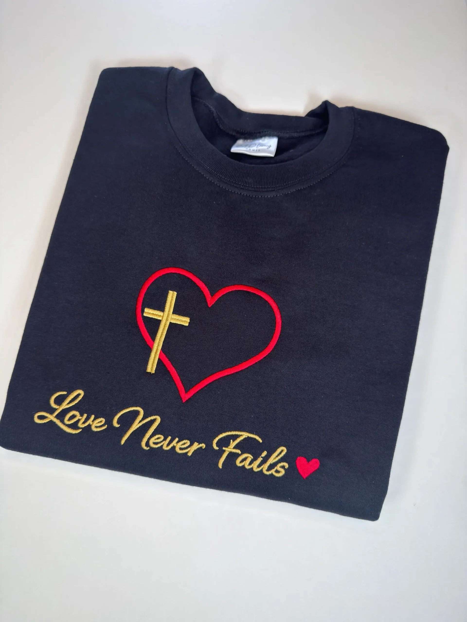 Black embroidered Christian T-shirt featuring red heart outline with gold cross and “Love Never Fails” Bible verse design